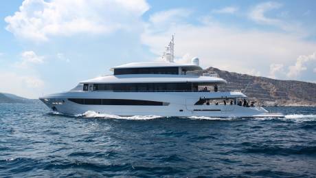 A sleek white yacht glides through the deep blue ocean waters near a rocky, mountainous coastline under a bright, cloudy sky.