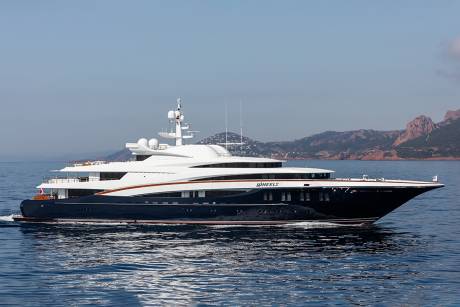 The luxurious yacht Wheels glides across the calm blue sea with a mountainous coastline in the background.
