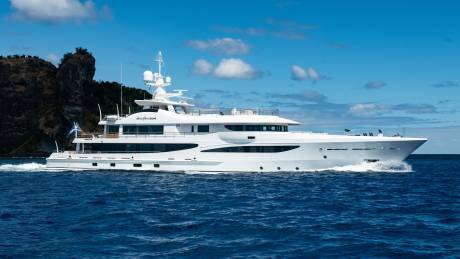 A luxurious white yacht glides through the deep blue ocean waters near a rocky, tree-covered island.