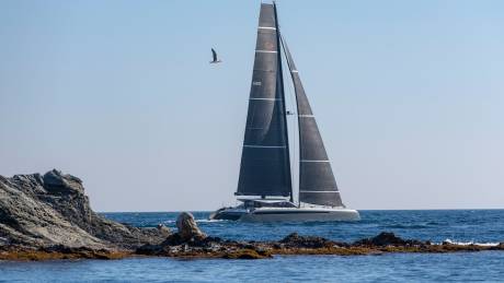 A sleek catamaran glides through the azure waters near a rocky coastline under a clear blue sky.