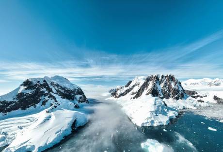 Towering snow-capped mountains emerge from a misty channel of icy water under a vast blue sky.