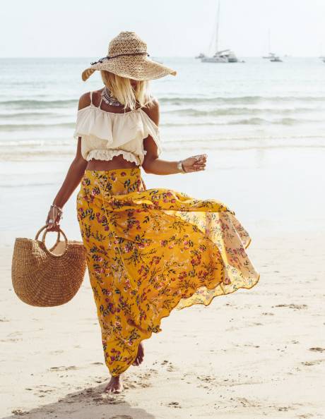 A woman in a flowy floral skirt and straw hat walks along a sandy beach with a woven bag in hand.