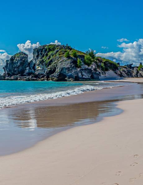 Turquoise waves gently lap a pink sand beach beneath dramatic rocky cliffs under a bright blue sky.