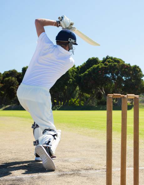 A cricketer in white pads and helmet swings his bat at the wicket on a sunny day.