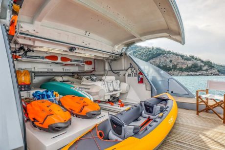 The image shows the interior of a boat or yacht, with various equipment and gear such as life jackets, bags, and other supplies neatly arranged on the deck. In the background, a scenic coastal landscape can be seen, suggesting the boat is situated in a picturesque location.