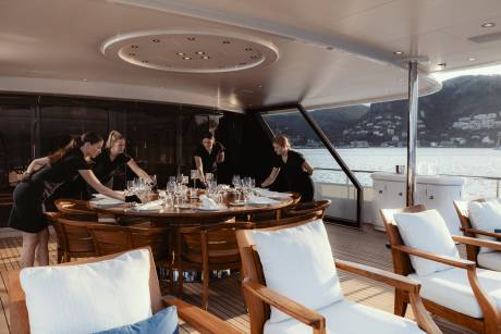 Four servers meticulously set a large dining table on a yacht's deck, overlooking a picturesque coastal town.