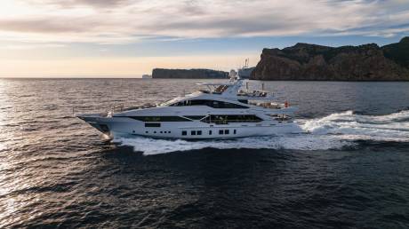 A sleek white yacht cuts through the dark blue sea, leaving a trail of white foam against a backdrop of rugged cliffs and a soft sunset sky.