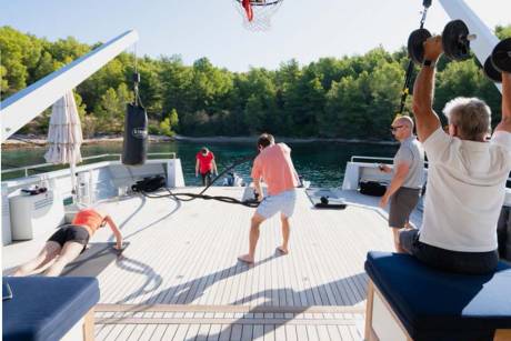 Four people engage in various exercises on the deck of a yacht anchored in a scenic, tree-lined bay.