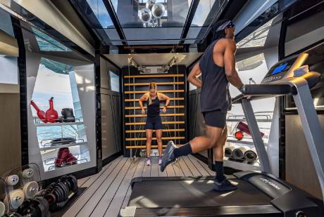A man runs on a treadmill while a woman uses resistance bands in a well-equipped gym on a luxurious yacht.