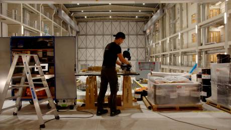 A worker in a cap and gloves operates a cutting saw near stacked containers in a vast, industrial warehouse.