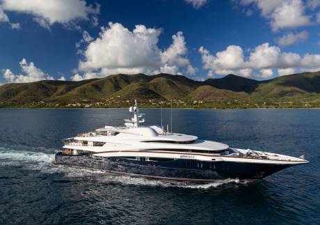 A luxurious yacht named Wheels glides through the azure waters with lush green hills in the background.
