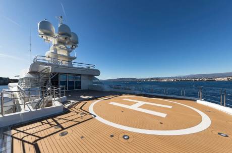 A luxurious superyacht's helipad offers stunning views of a coastal city under a clear blue sky.