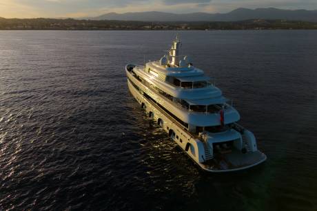 A luxurious white yacht glides across calm waters as the sun sets behind a distant coastal town.