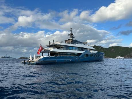 A large, luxurious yacht with a sleek, modern design sits in the calm, blue waters, surrounded by lush, green hills and a picturesque cloudy sky.
