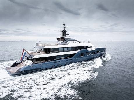 A large, sleek yacht with multiple decks and a tall mast is speeding across the choppy, gray waters of what appears to be an open sea or lake, with a cloudy, overcast sky in the background.