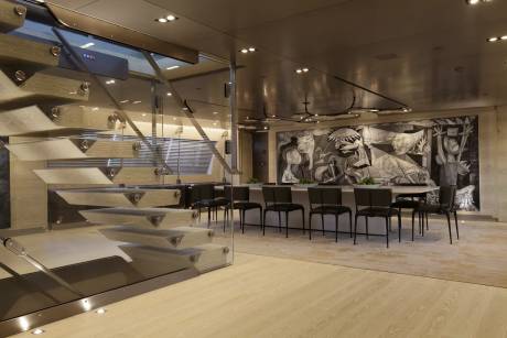 A modern dining room features a striking mural above a long table with minimalist chairs, complemented by a sleek glass staircase.