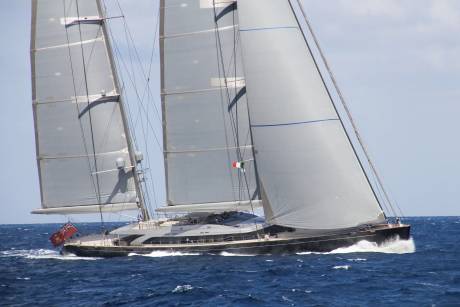 A sleek black yacht with towering sails cuts through the deep blue ocean under a clear sky.