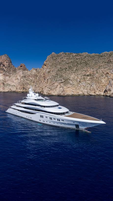 A magnificent white superyacht glides through deep blue waters near a rugged, rocky coastline under a clear sky.