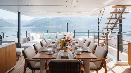 A beautifully set dining table awaits guests on a luxurious yacht deck, overlooking a serene bay with verdant mountains.