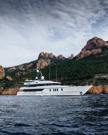 A luxurious white yacht cruises serenely past rugged, tree-covered mountains under a dramatic sky.