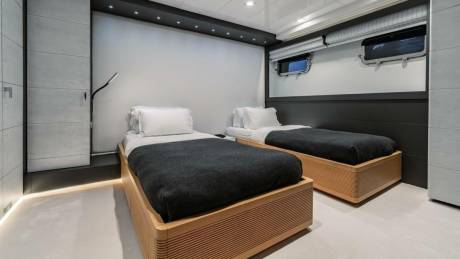 Two twin beds with dark gray blankets and white pillows are neatly made in a modern cabin with porthole windows.