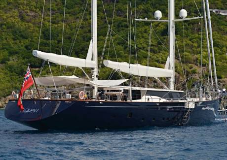 A majestic dark blue sailing yacht glides through calm blue waters with lush green hills in the background.