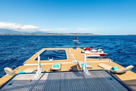 A person on a jet ski glides across the sparkling blue ocean near a luxurious yacht with a pool and lounge chairs.