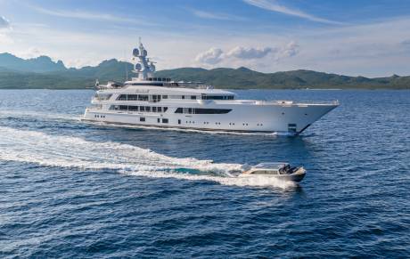 A luxurious superyacht cruises through the azure waters, leaving a frothy wake behind it, with a smaller tender boat in tow.