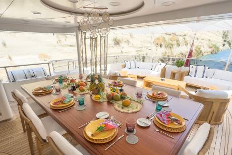 A beautifully set dining table on a yacht deck awaits a meal with a scenic backdrop of cliffs and water.