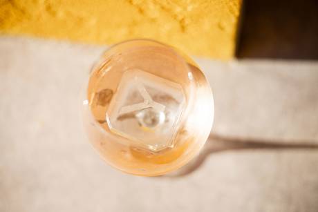 A close-up overhead view captures a glass bottle filled with a golden liquid, casting a soft shadow on a textured surface.