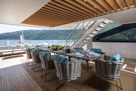 An elegant outdoor dining area on a yacht features a long table set for a meal with a backdrop of lush green hills and sparkling blue water.