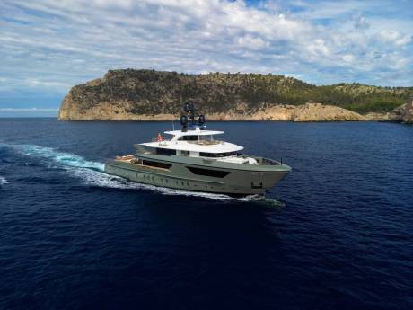 A sleek, modern yacht glides through the deep blue waters near a rugged, tree-covered island under a cloudy sky.