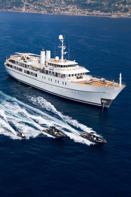 A luxurious yacht cruises through the deep blue sea, accompanied by two smaller tenders creating white wakes.