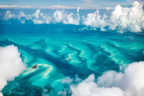 Aerial view of turquoise waters revealing intricate sandbars and scattered islands beneath fluffy white clouds.