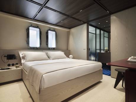 A modern bedroom features a light wood bed and minimalist decor, with porthole-style windows offering a glimpse of the outside.