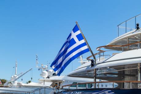 A large white yacht with a Greek flag flying proudly on its stern, set against a clear blue sky.