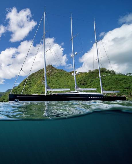 A sleek black schooner with tall masts glides through clear turquoise waters near a lush green island.