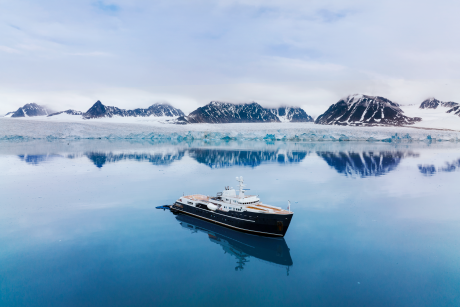 A sleek, dark-colored yacht floats peacefully in the serene, mirror-like waters, surrounded by the majestic, snow-capped mountains of the Arctic landscape.
