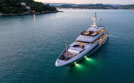 A large, luxurious yacht is anchored in a serene, turquoise bay surrounded by lush, verdant hills and buildings in the distance.