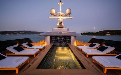 A luxurious yacht with a sleek design, featuring a central structure with a cross on top, surrounded by lounge chairs and a pool-like area, set against a scenic backdrop of a body of water and a night sky.