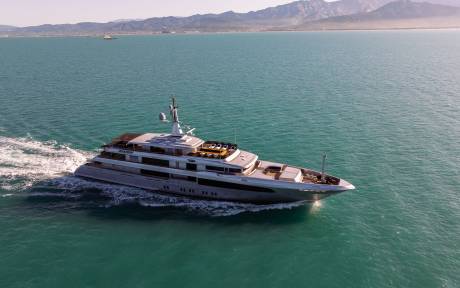 A large, luxurious yacht is speeding across a serene, turquoise-colored body of water, with a mountainous landscape visible in the background.