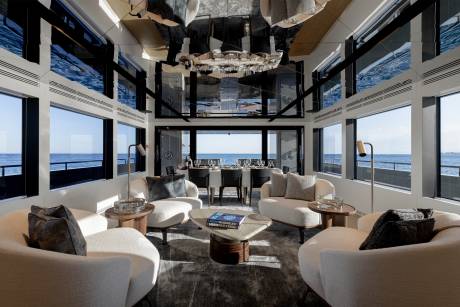 A luxurious and spacious living room with large windows offering a stunning view of the ocean, featuring plush white sofas, a grand chandelier, and a dining area in the background.