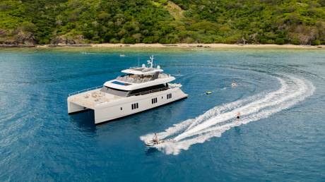 A sleek catamaran yacht floats on calm blue water in the foreground, while a person on a jet ski creates a white wake, and a lush green, tree-covered island rises in the background.
