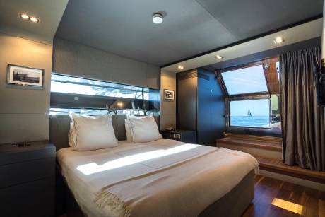 A neatly made bed with white pillows dominates the foreground, while a large window in the background offers a view of the ocean and a distant sailboat.