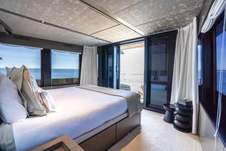 A luxurious bedroom with a large bed in the foreground opens to a balcony overlooking the ocean in the background.