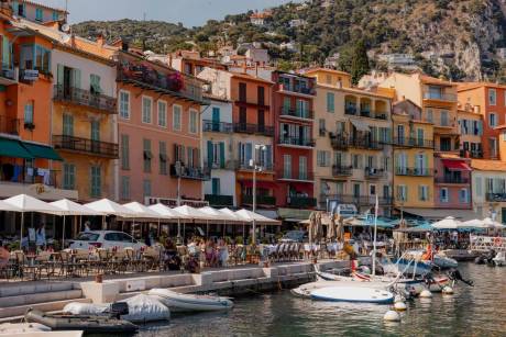 Colorful buildings line a waterfront promenade with outdoor cafes in the foreground, while a lush, green hillside rises behind them.