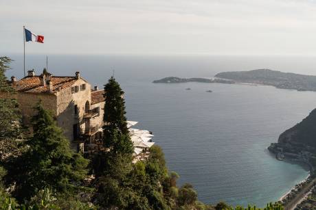 A stone building with a French flag flies on a cliff overlooking a calm blue sea with islands in the distance.