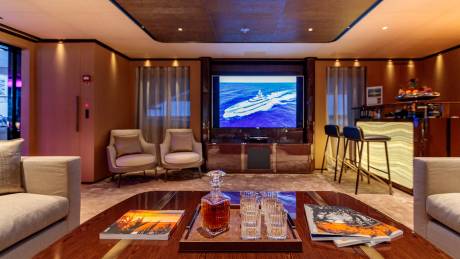 A decanter of whiskey and glasses sit on a coffee table in the foreground, while a large television displays a ship at sea in the background.