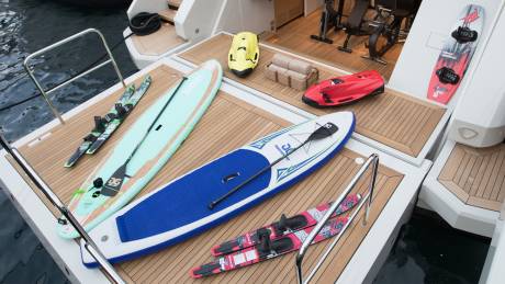 In the foreground, paddleboards and skis rest on a yacht's deck, while the background reveals exercise equipment and watercraft.