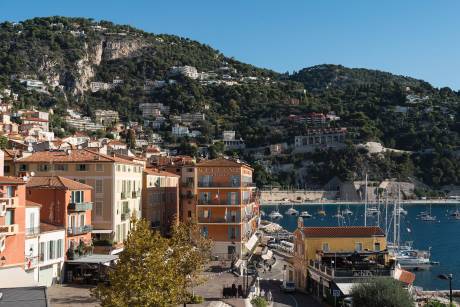 Colorful buildings line a waterfront street in the foreground, with a bay dotted with sailboats and a lush, green hillside dotted with more buildings in the background.